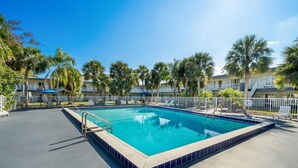 A rectangular pool with a tiled edge, surrounded by a white fence and palm trees. - Suburban Studios Daytona Beach - Speedway (Daytona Beach)