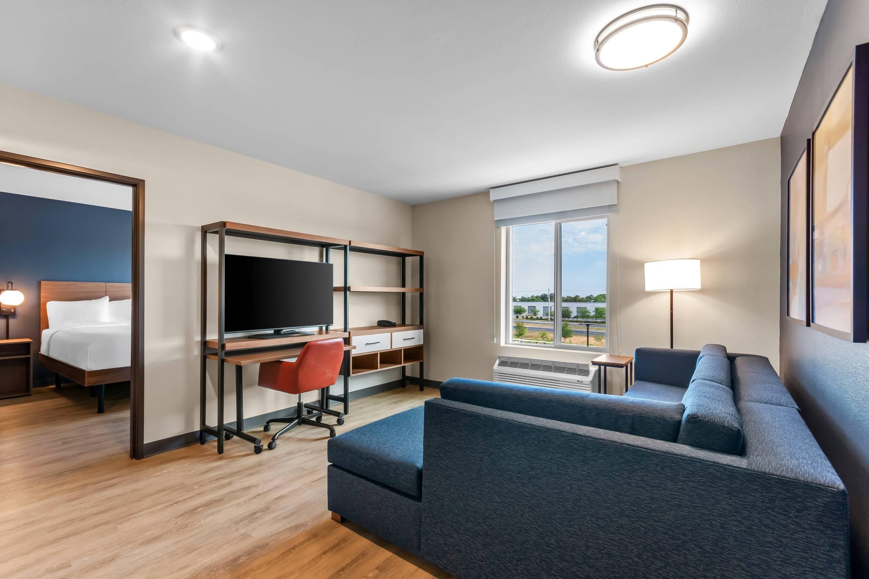 A hotel room with a bed, a desk with a computer, a sofa, a TV, and a window with a view.