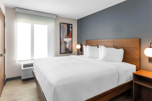 A hotel room with a large bed, a nightstand, a wall-mounted lamp, a window, and a radiator. - Everhome Suites Panama City Beach - 30A (Panama City Beach)