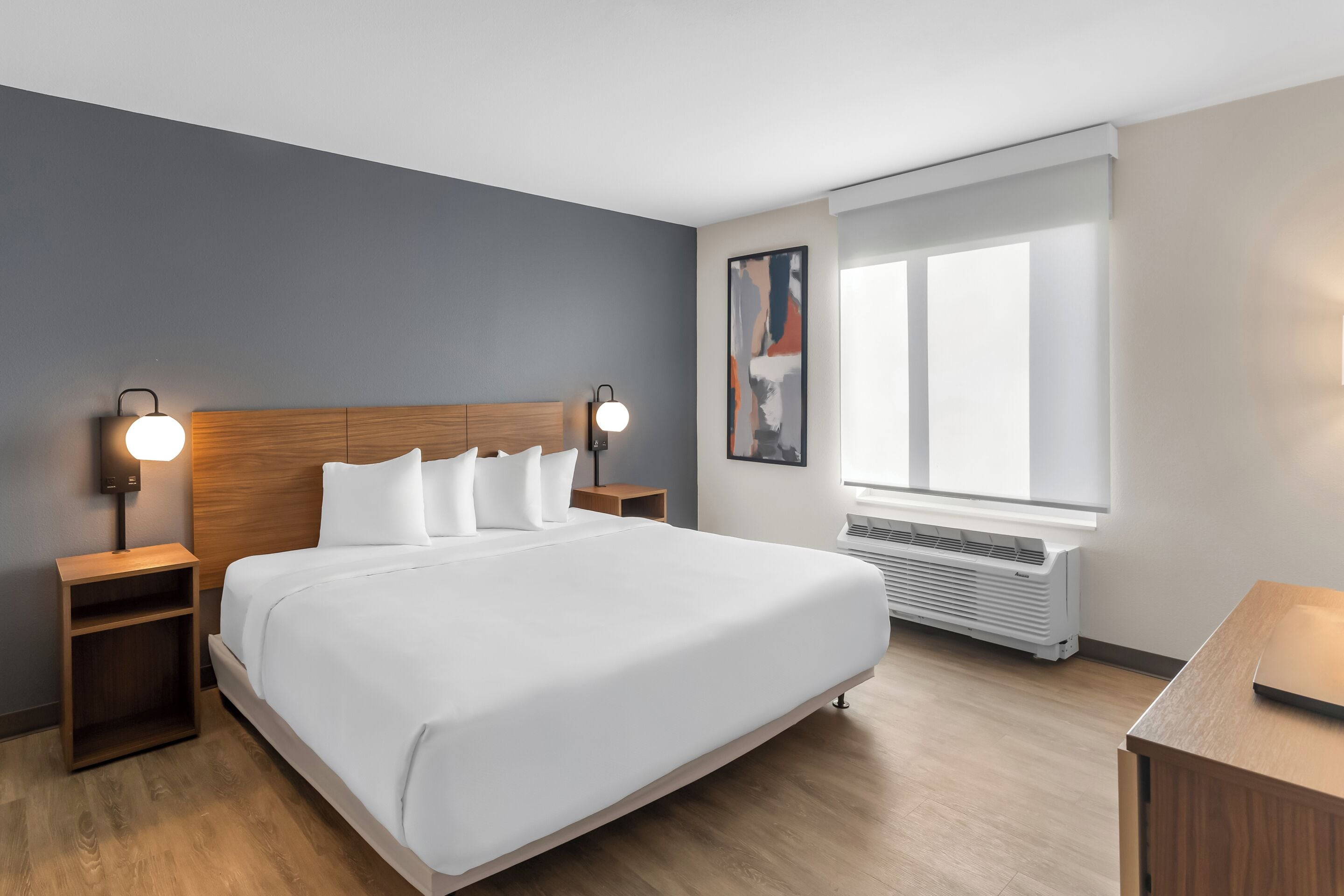 Una habitación de hotel moderna con una cama grande, mesitas de noche, un escritorio, una ventana con persianas y aire acondicionado instalado en la pared.