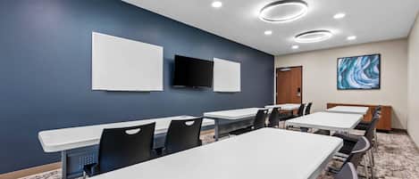 Un salón de conferencias con mesas y sillas blancas, un televisor de pantalla plana y un cuadro en la pared.