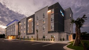 A multi-story hotel with a modern design, featuring a prominent 'EveryHome Suites' sign, a parking lot, and a well-maintained lawn with a palm tree.