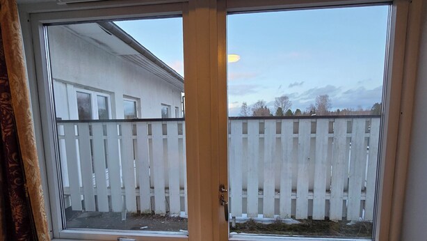 A room with a window and a door, a white wall, and a view of a white fence and trees. - Hotell Våler-Elverum-flisa-kirkenær (Våler)