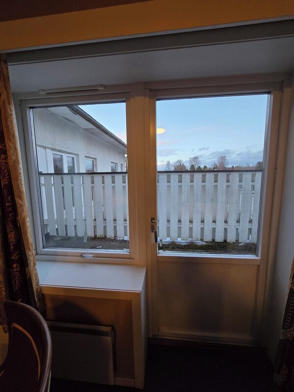 A room with a window and a door, a white wall, and a view of a white fence and trees. - Hotell Våler-Elverum-flisa-kirkenær (Våler)