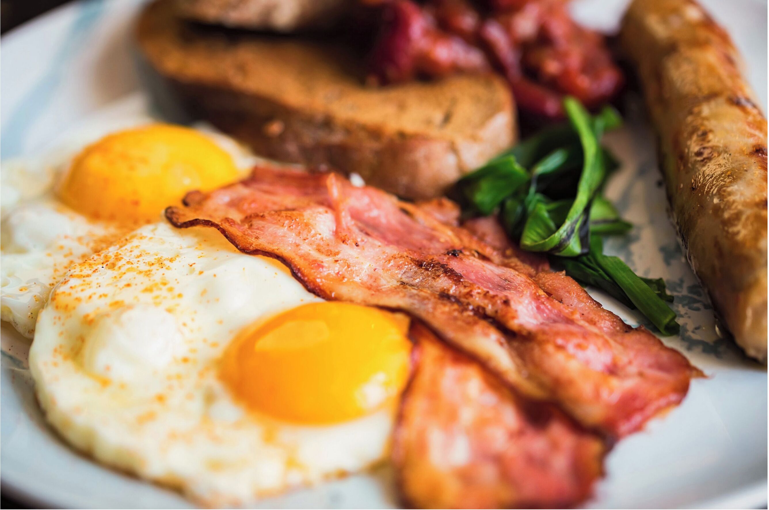 En morgenmadstallerken med æg, bacon, pølser og grønt.