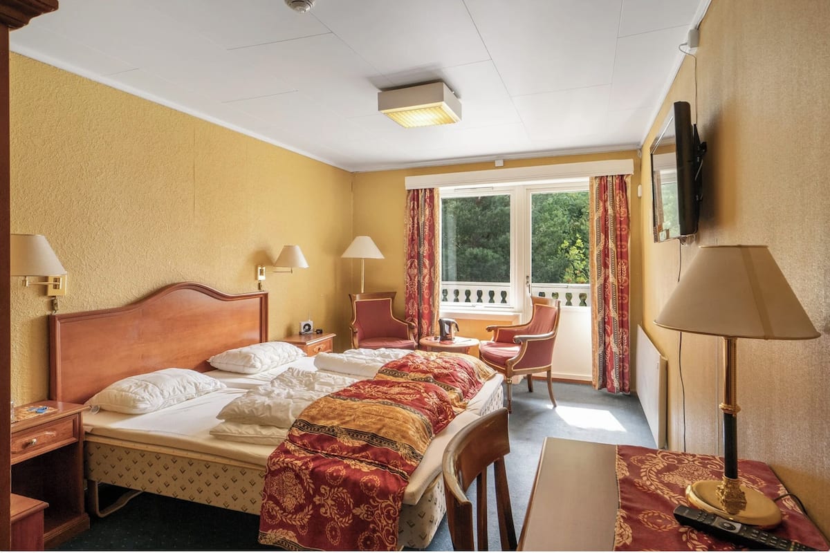 A hotel room with a bed, two chairs, a desk, a TV, and a window with curtains.