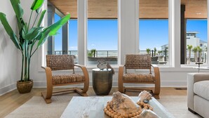 A modern living room with large windows, two wicker chairs, a coffee table with decorative shells, and a potted plant.