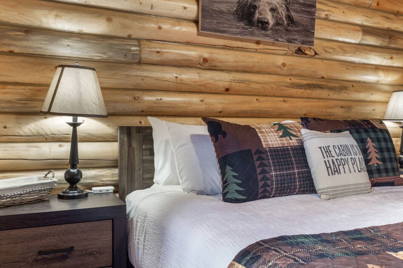 A cozy cabin-style room with a wooden log wall, a bed with patterned pillows, and a lamp on a nightstand.