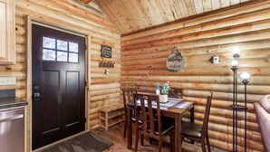 A log cabin interior with a dark wooden door, a dining table with chairs, a small bench, a potted plant, and a wall-mounted light fixture. - The Bear`s Burrow | 7 Mi to YNP | Private Pond (West Yellowstone)