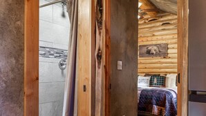 A wooden cabin interior with a shower, a bed with a plaid blanket, and a wooden ceiling. - The Bear`s Burrow | 7 Mi to YNP | Private Pond (West Yellowstone)