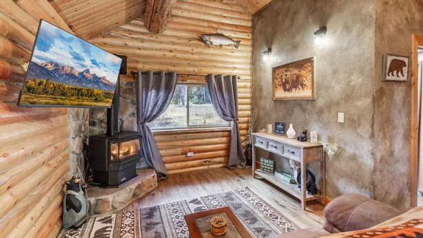 A log cabin interior with a stone fireplace, a television, a sofa, and a window overlooking a forested area. - The Bear`s Burrow | 7 Mi to YNP | Private Pond (West Yellowstone)