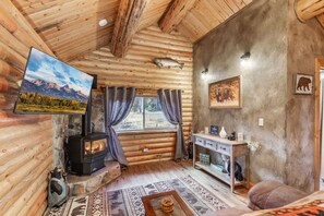 A log cabin interior with a stone fireplace, a television, a sofa, and a window overlooking a forested area. - The Bear`s Burrow | 7 Mi to YNP | Private Pond (West Yellowstone)