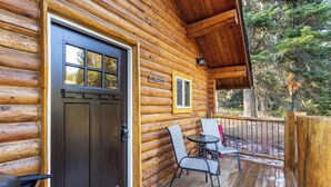 A log cabin with a dark wooden door, a welcome sign, a small table with two chairs, and a deck with a railing. - The Bear`s Burrow | 7 Mi to YNP | Private Pond (West Yellowstone)