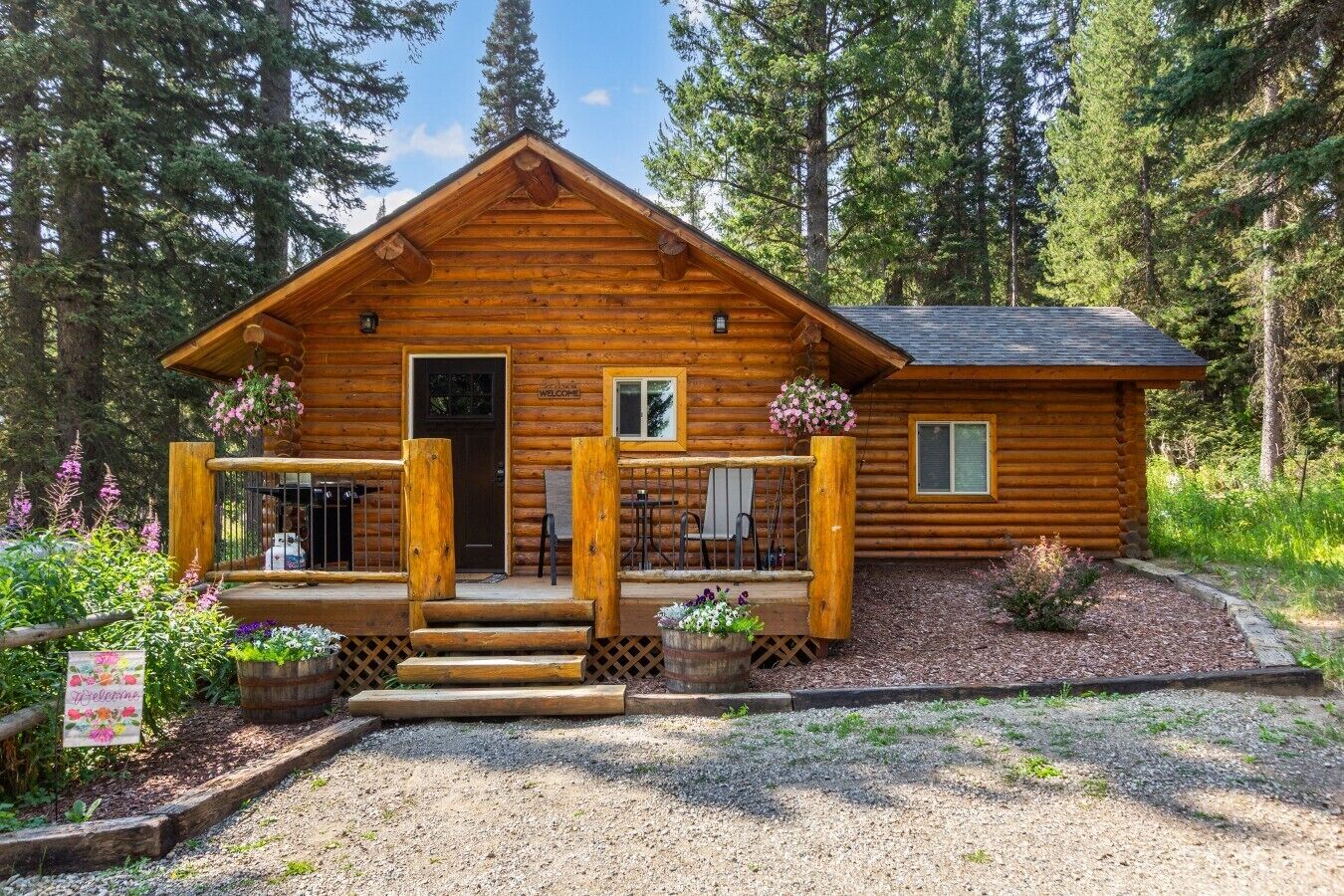 A wooden cabin with a porch, a welcome sign, and a gravel driveway.