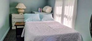 A bedroom with a bed, a nightstand, a lamp, a small table, and a window with curtains. - Welcome to Bluebird Cottage (Lakeview)