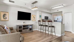 Ein modernes Wohnzimmer mit Flachbildfernseher, einer Couch, einem Couchtisch und einer Küchenzeile inklusive Mikrowelle, Kühlschrank und Barhocker.