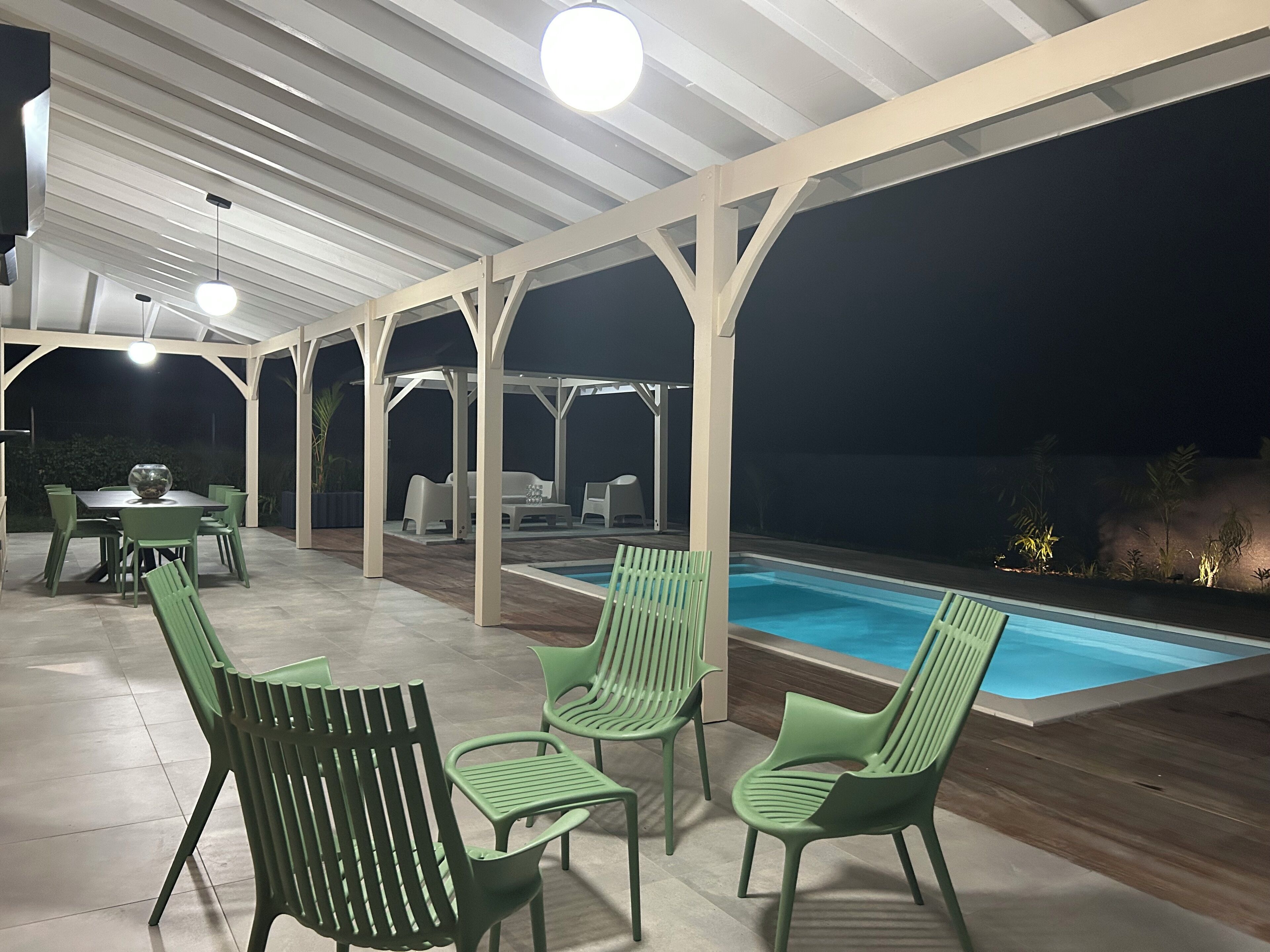 An outdoor patio area with a pool, green chairs, a dining table, and a covered seating area.