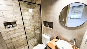 A modern bathroom with a glass-enclosed shower, a toilet, a sink with a wooden vanity, and a round mirror on the wall. - Rent Up - Estudio Moderno en Palermo (Buenos Aires)