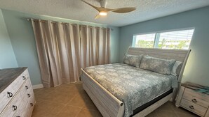 Ein Schlafzimmer mit Bett, Schminktisch, Nachttisch, Deckenventilator und Fenster mit Jalousien.