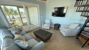 Ein Wohnzimmer mit grauem Sofa, gestreifte Sessel, einem gewebten Couchtisch und einem Fernseher an der Wand.