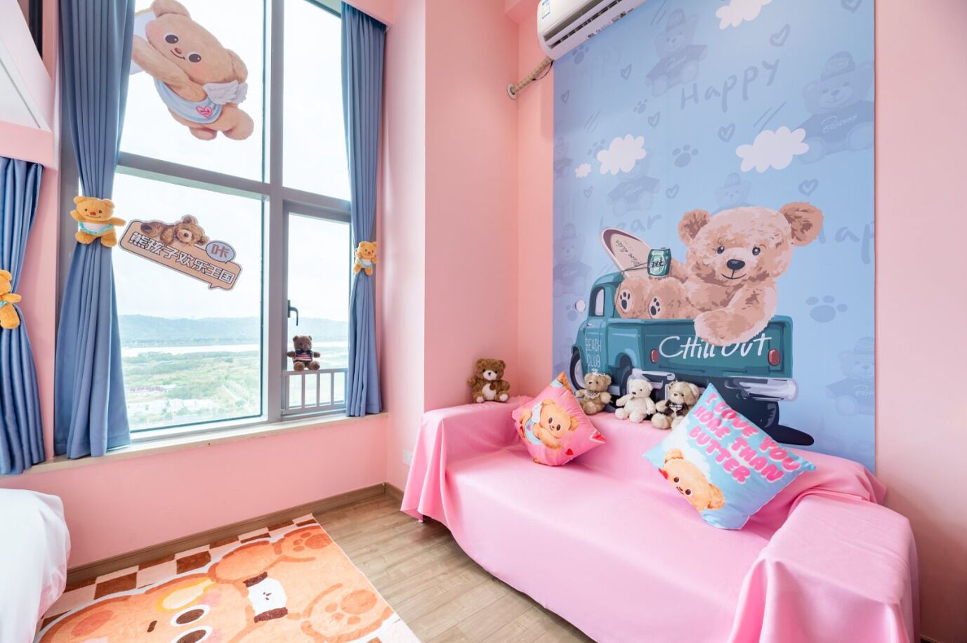 Een kamer met een roze thema, met een grote muurschildering van een teddybeer, een roze bank en uitzicht op een landschap door het raam.