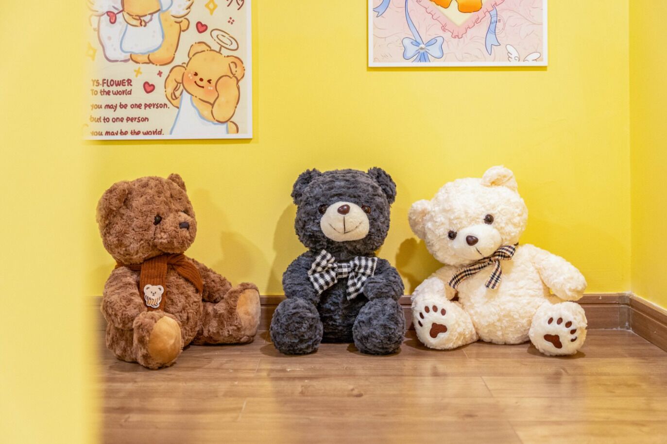 Three teddy bears sitting on a wooden floor against a yellow wall with two framed pictures above them.