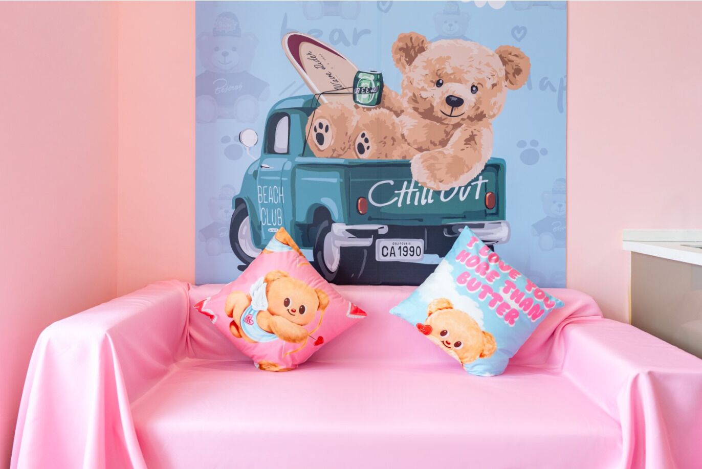 A pink-themed room with a bed, two pillows, and a wall mural of a teddy bear on a vintage car.