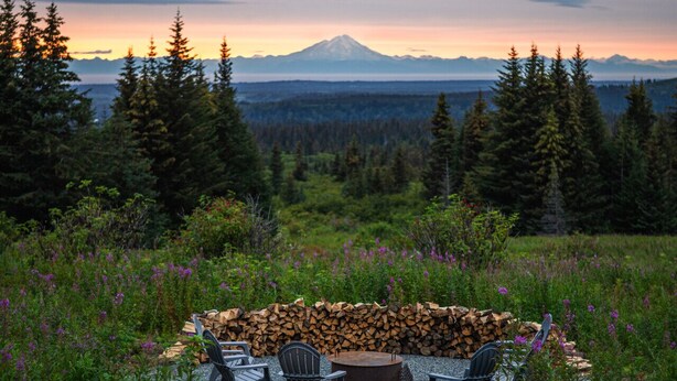 An outdoor seating area with a gravel path leading to a table and chairs, surrounded by greenery and a mountain view. - Misty Ridge (Homer)