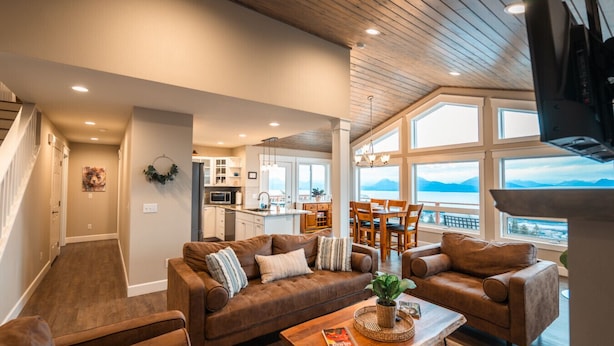 A spacious living room with a large brown leather sofa set, a wooden coffee table, and a view of a kitchen and dining area through large windows. - Little Bayview Cottage (Homer)