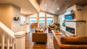 A spacious living room with a fireplace, wooden ceiling, and large windows overlooking a scenic view. - Little Bayview Cottage (Homer)