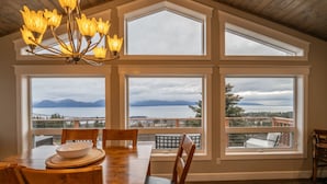 A dining area with a large table and chairs, large windows with a view of mountains and a lake, and a chandelier. - Little Bayview Cottage (Homer)