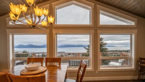 A dining area with a large table and chairs, large windows with a view of mountains and a lake, and a chandelier. - Bayview Cottage (Homer)
