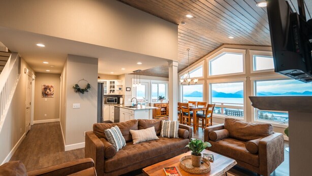 A spacious living room with a large brown leather sofa set, a wooden coffee table, and a view of a kitchen and dining area through large windows. - Bayview Cottage (Homer)