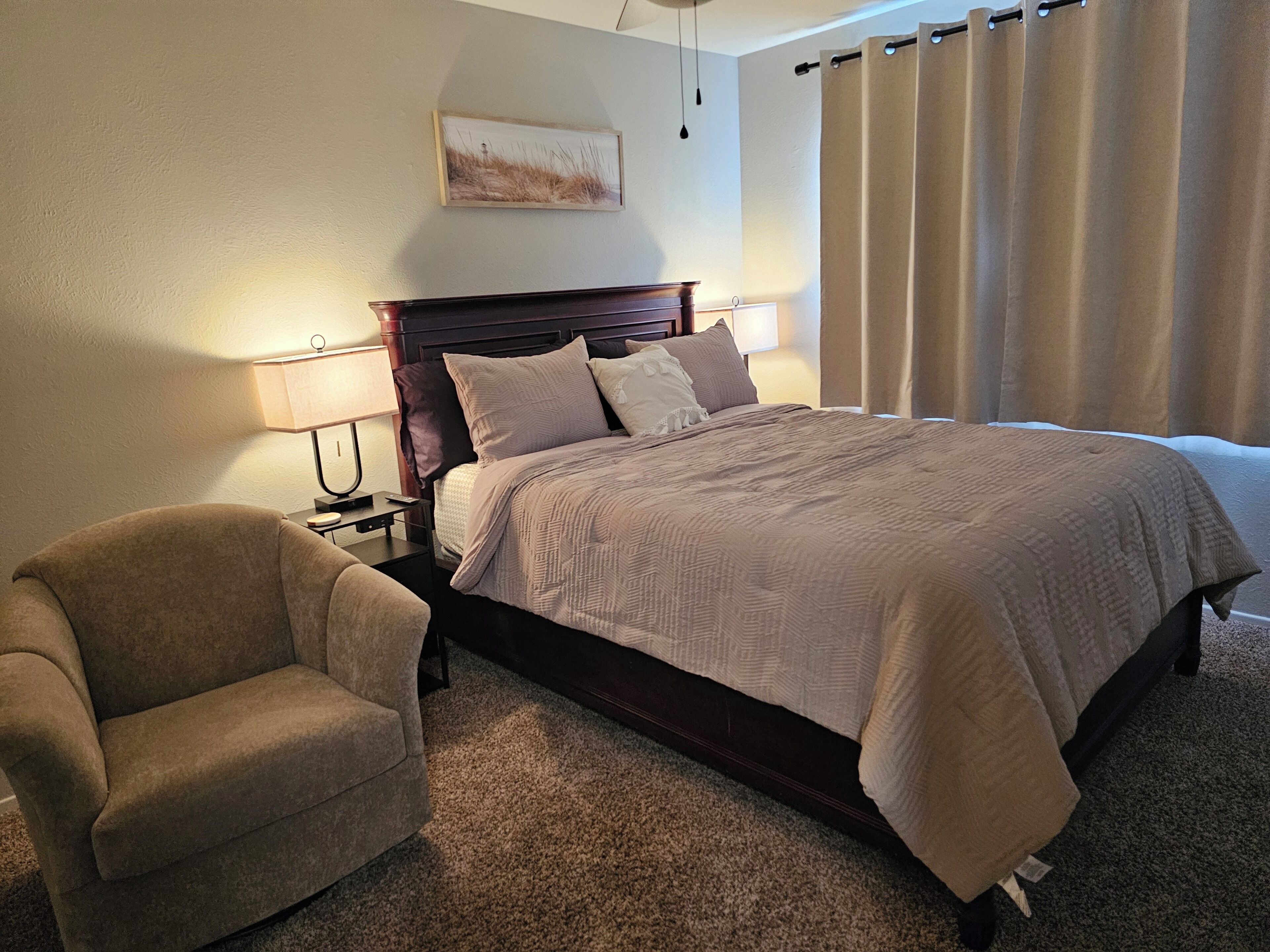 A hotel room with a bed, a chair, a nightstand, a lamp, a painting on the wall, and a window with curtains.