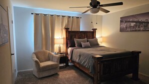 Een slaapkamer met een houten bed, een stoel, een plafondventilator en een nachtkastje met een lamp.