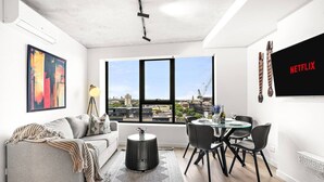 Een moderne woonkamer met een grijze bank, een eettafel met stoelen, een groot raam met uitzicht op de stad en een tv aan de muur waarop 