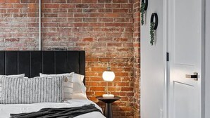 Une chambre avec un mur en briques, un lit avec une literie blanche, une table de chevet avec une lampe et une porte avec une poignée.