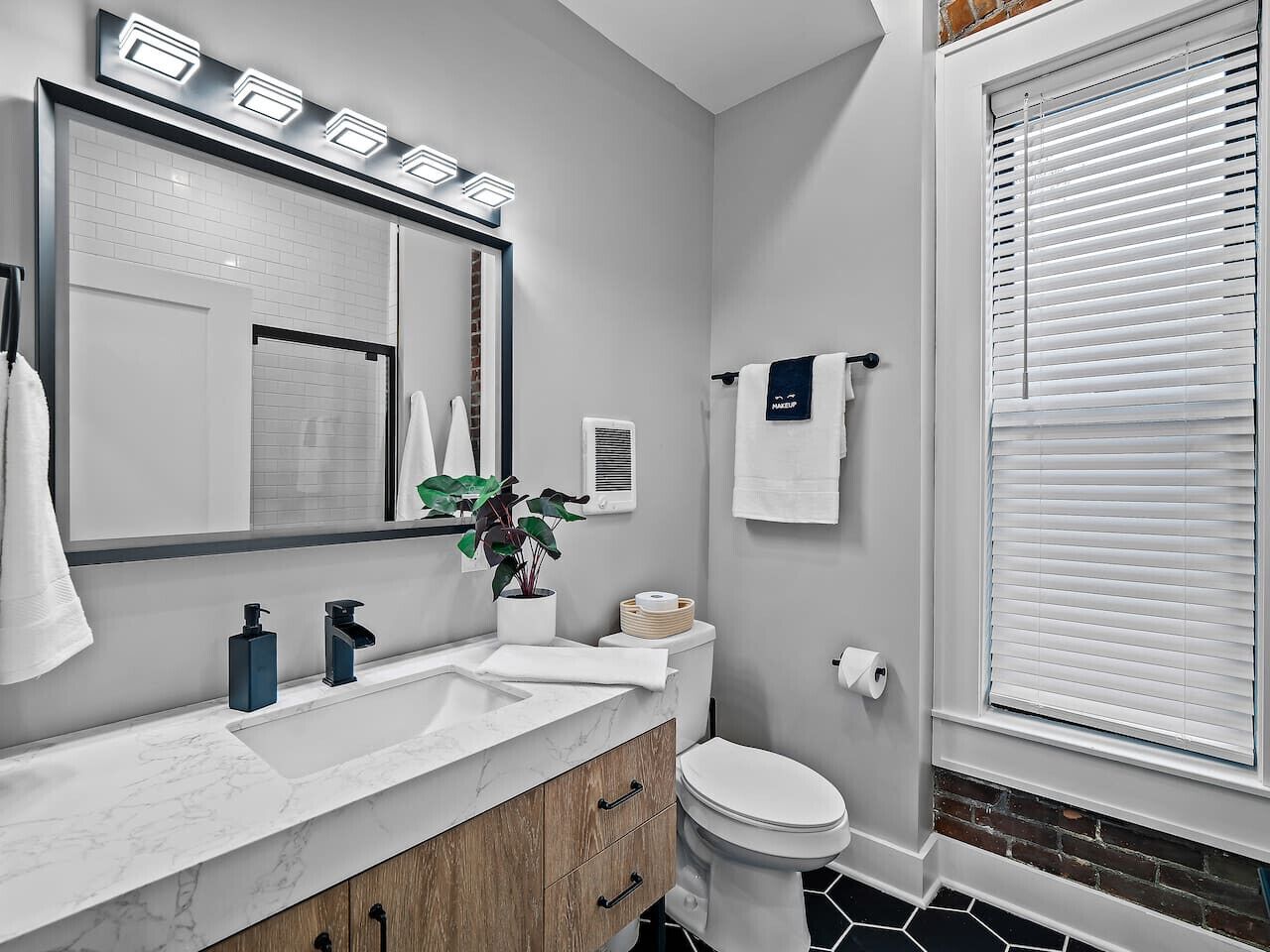 A modern bathroom with a large mirror, white sink, and toilet. There is a plant on the sink and a window with blinds.