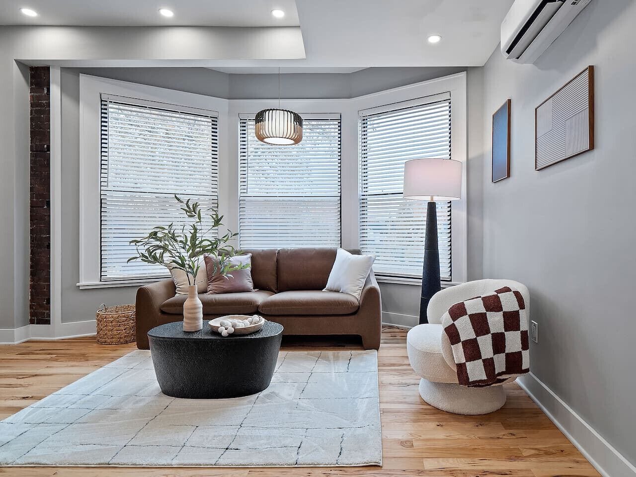 Una sala moderna con un sofá marrón, una mesa redonda de centro con una planta, un sillón a cuadros y una lámpara.