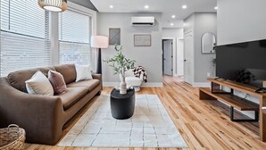 Un salon moderne avec un canapé, une table basse, un téléviseur et une grande fenêtre équipée de stores.