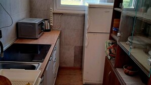 Een compacte keuken met een magnetron, koelkast en wastafel. Een raam zorgt voor daglicht en uitzicht op bomen.