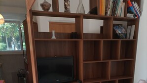 Een slaapkamer met een bed, een televisie, houten planken met boeken en een raam.