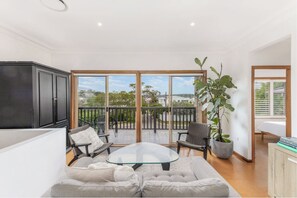 A modern living room with a glass coffee table, grey sofa, and black chairs. A large black cabinet is on the left, and a potted plant is on the right. - Bayside Retreat (Burraneer)