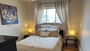 A bedroom with a bed, two bedside tables with lamps, a painting on the wall, and a window with curtains. - Furnished monthly rental Near Downtown Denver (Denver)