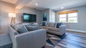 Ein modernes Wohnzimmer mit grauem Sofa, einem Flachbildfernseher und einem großen Fenster mit Jalousien.