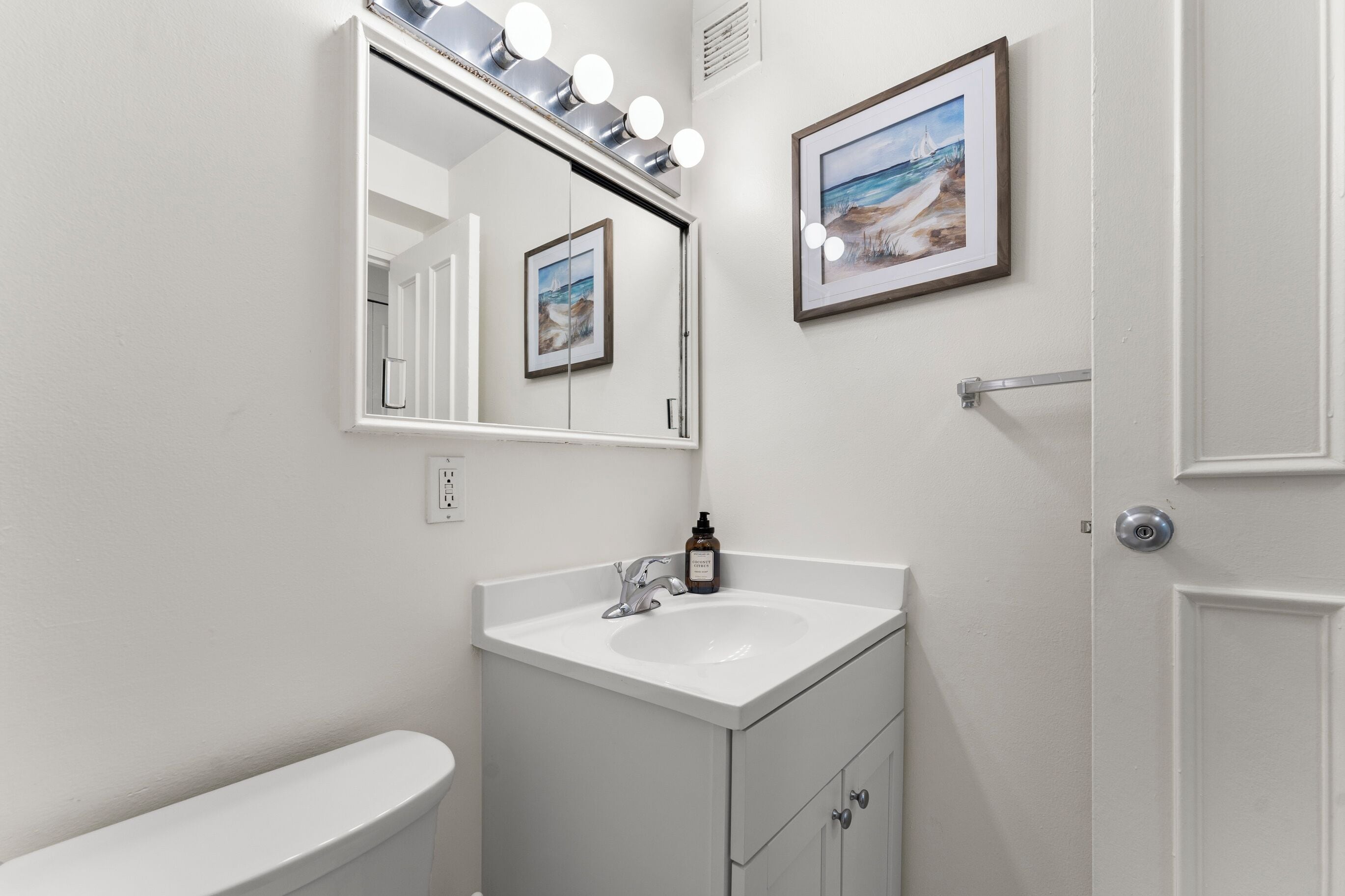 A bathroom with a white sink, a mirror with lights, and two framed pictures on the wall.