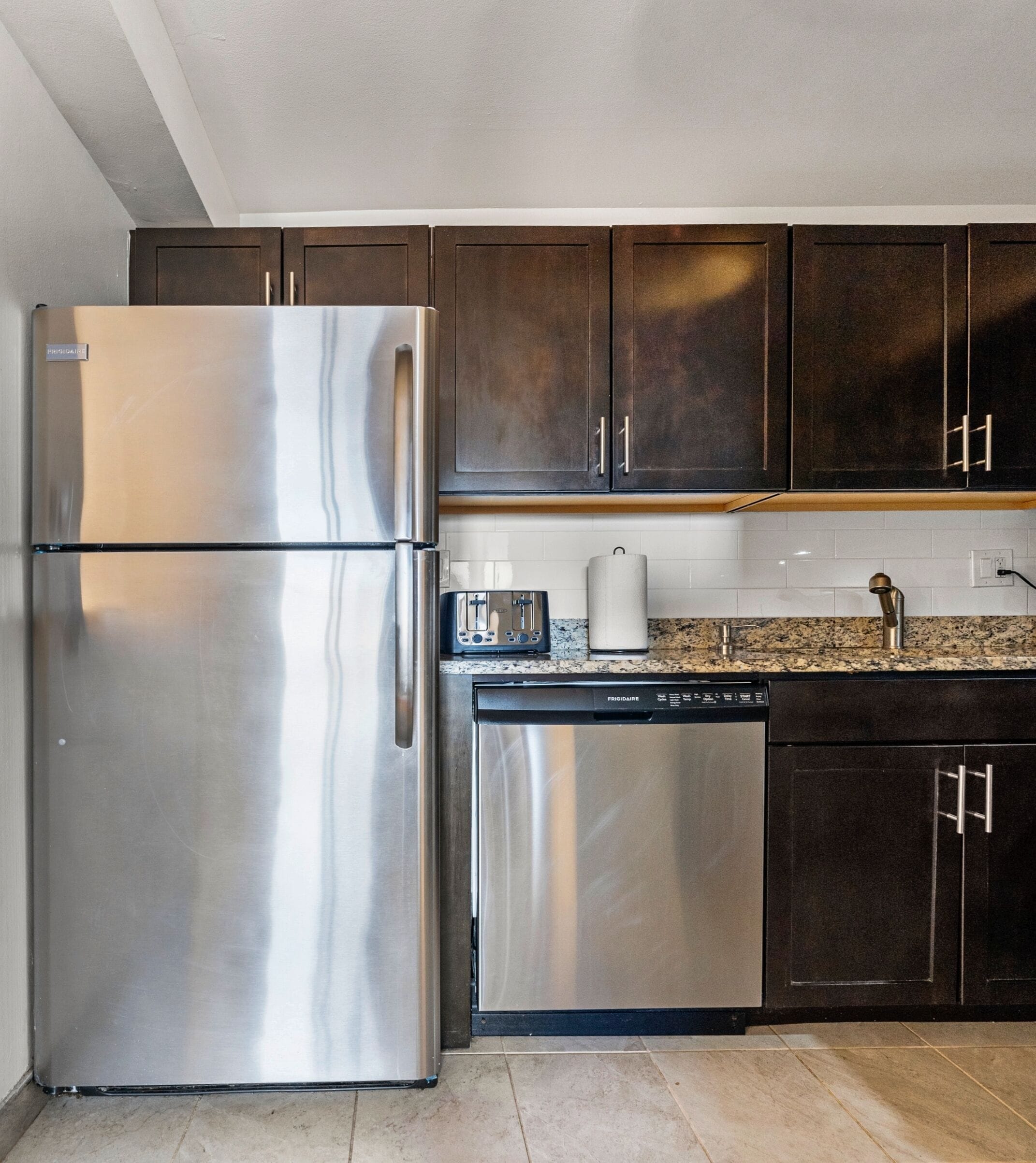 Una cocina con refrigerador de acero inoxidable, lavavajillas, armarios de madera oscura, encimera de granito y salpicadero de azulejos blancos.