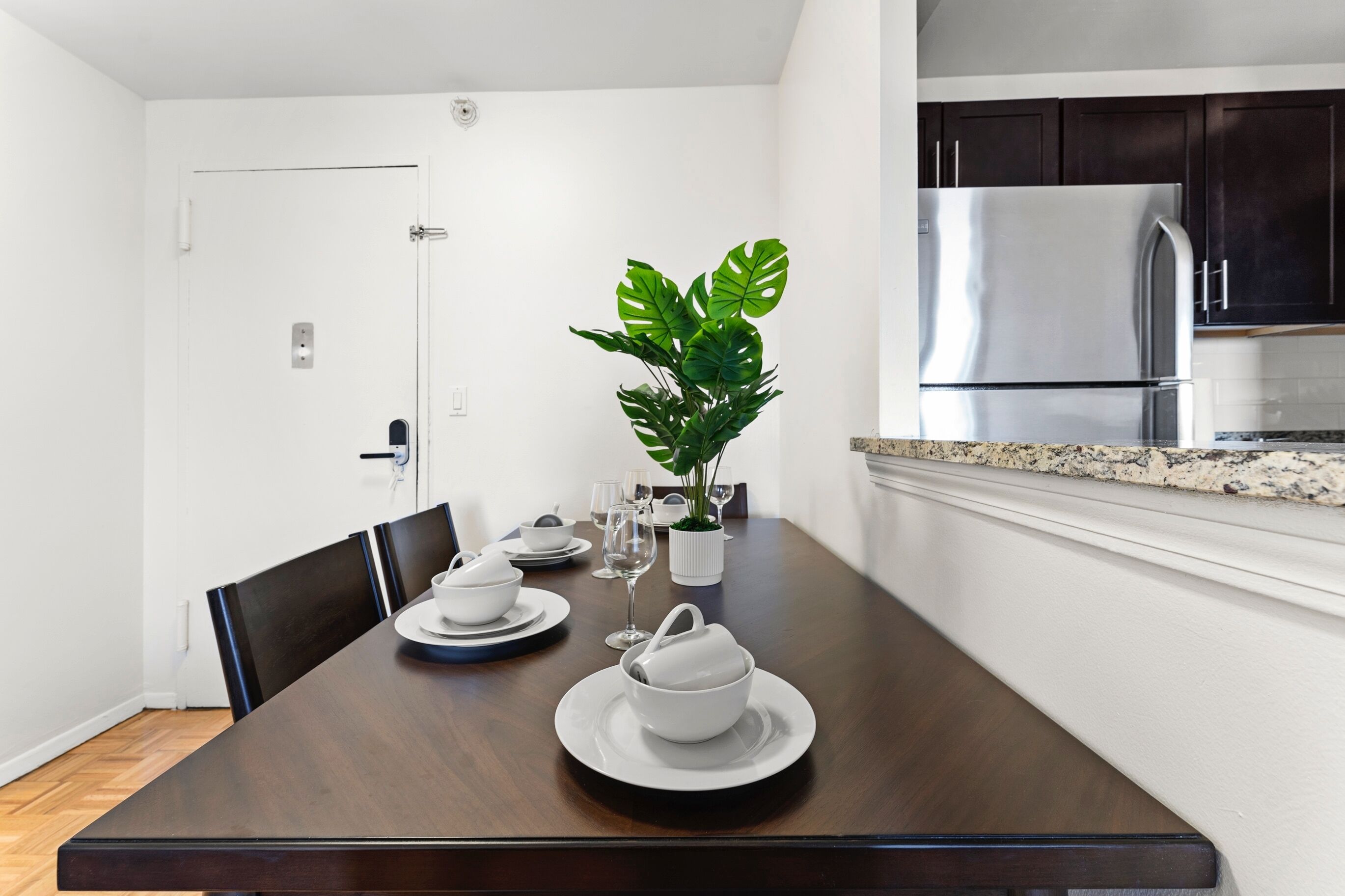 A dining area with a dark wooden table set for four, white plates, bowls, and glasses. A potted plant is on the table, and a kitchen with dark cabinets and stainless steel appliances is visible in the background.