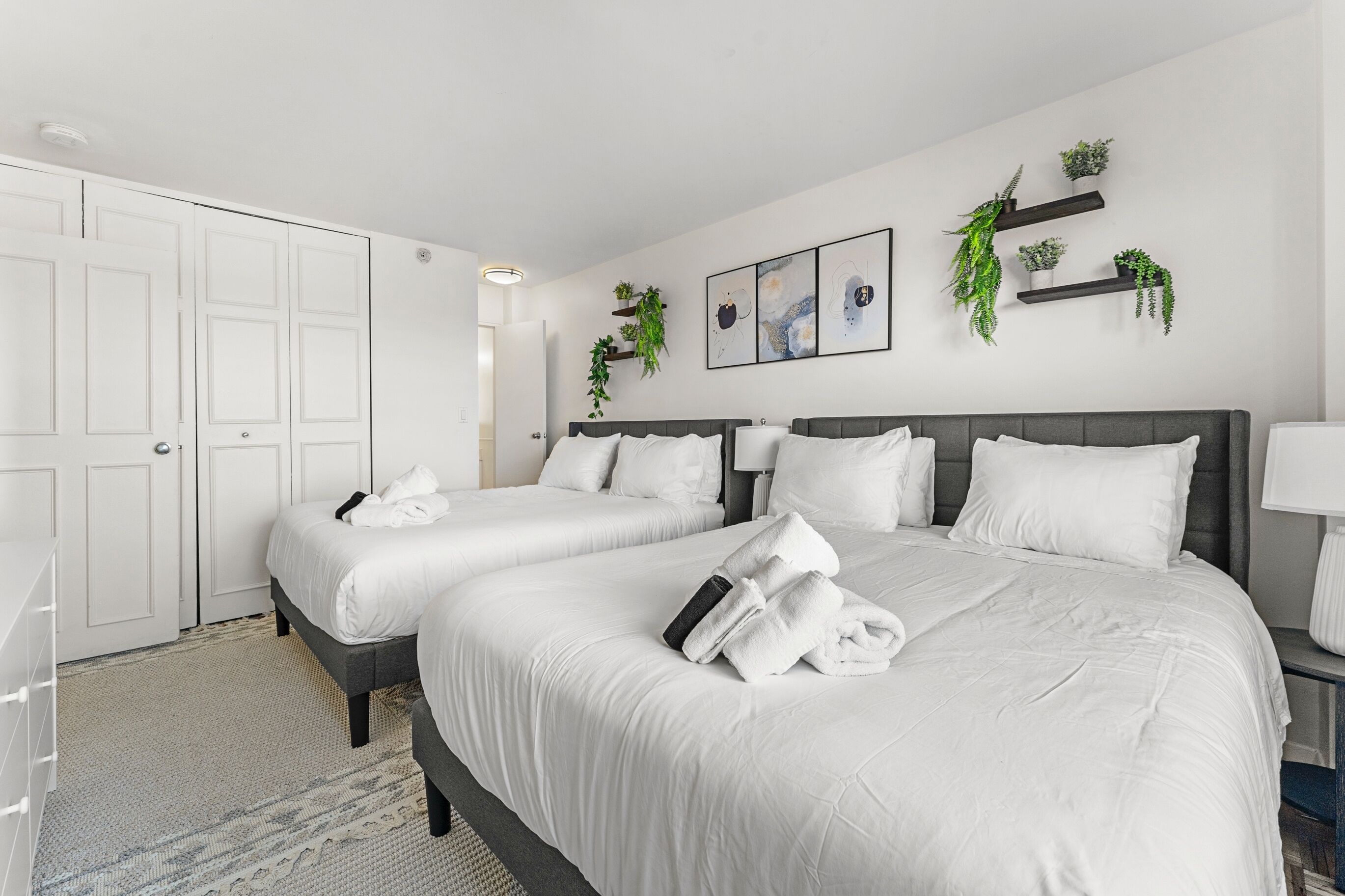 A hotel room with two beds, white bedding, a gray headboard, a bedside table with a lamp, and wall-mounted shelves with plants.