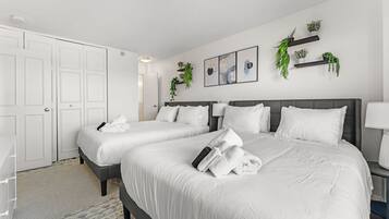A hotel room with two beds, white bedding, a gray headboard, a bedside table with a lamp, and wall-mounted shelves with plants.